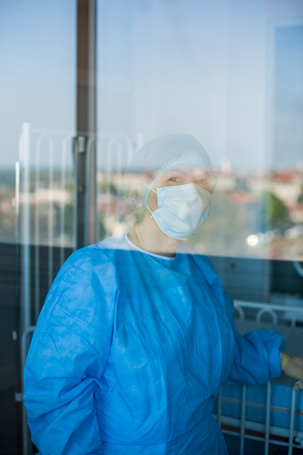 Une infirmière dans une salle stérile pose à travers la vitre. On voit Toulouse dans le reflet de la vitre
